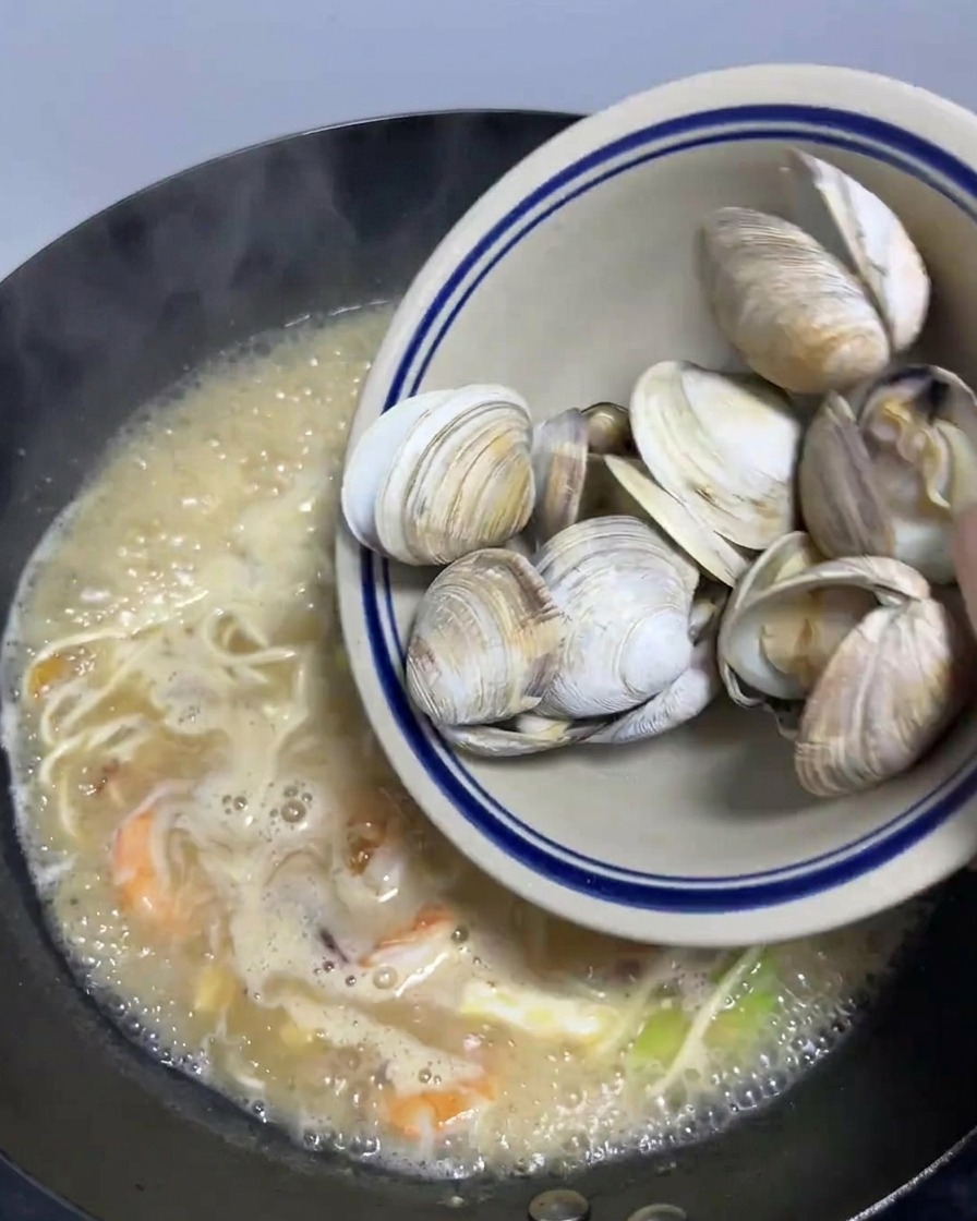 A bowl of blanched clams being poured into a large wok filled with simmering seafood ramen.