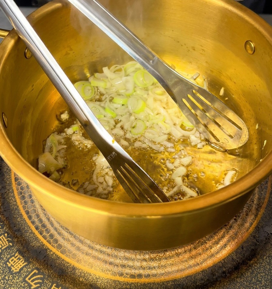 Raw pink minced pork being stirred into sautéed garlic and scallions inside a golden pot using metal tongs.