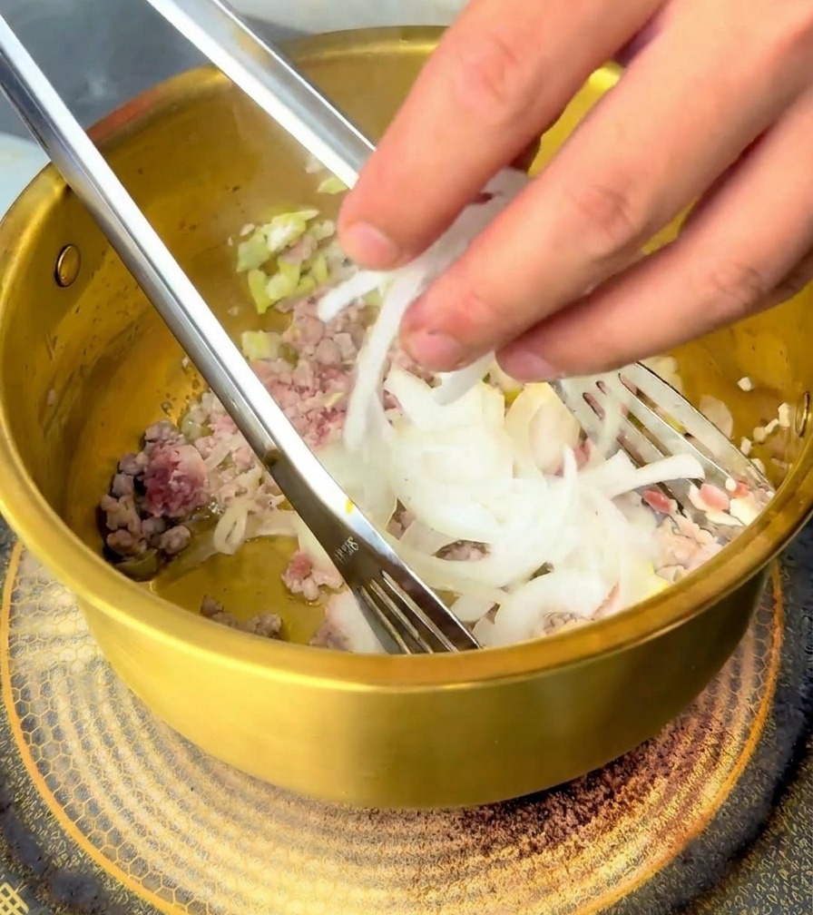 A hand adding a handful of sliced white onions into a golden brass pot containing cooked minced meat and aromatics.