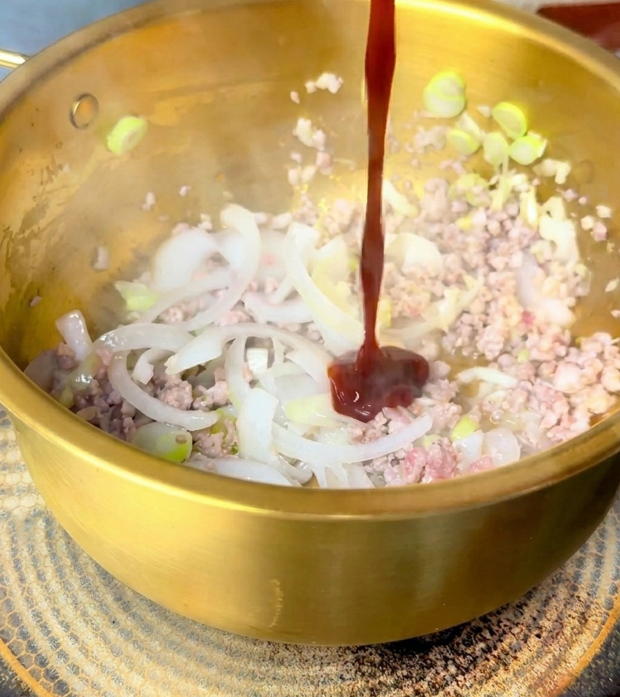 A thick stream of dark red sauce being poured over stir-fried pork and onions in a golden pot.