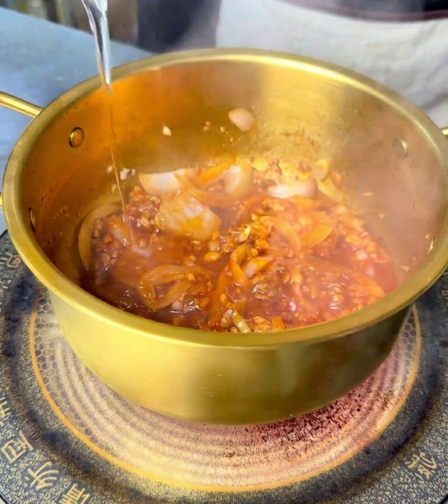 Clear water being poured into a brass pot, mixing with a red spicy base and stir-fried ingredients.
