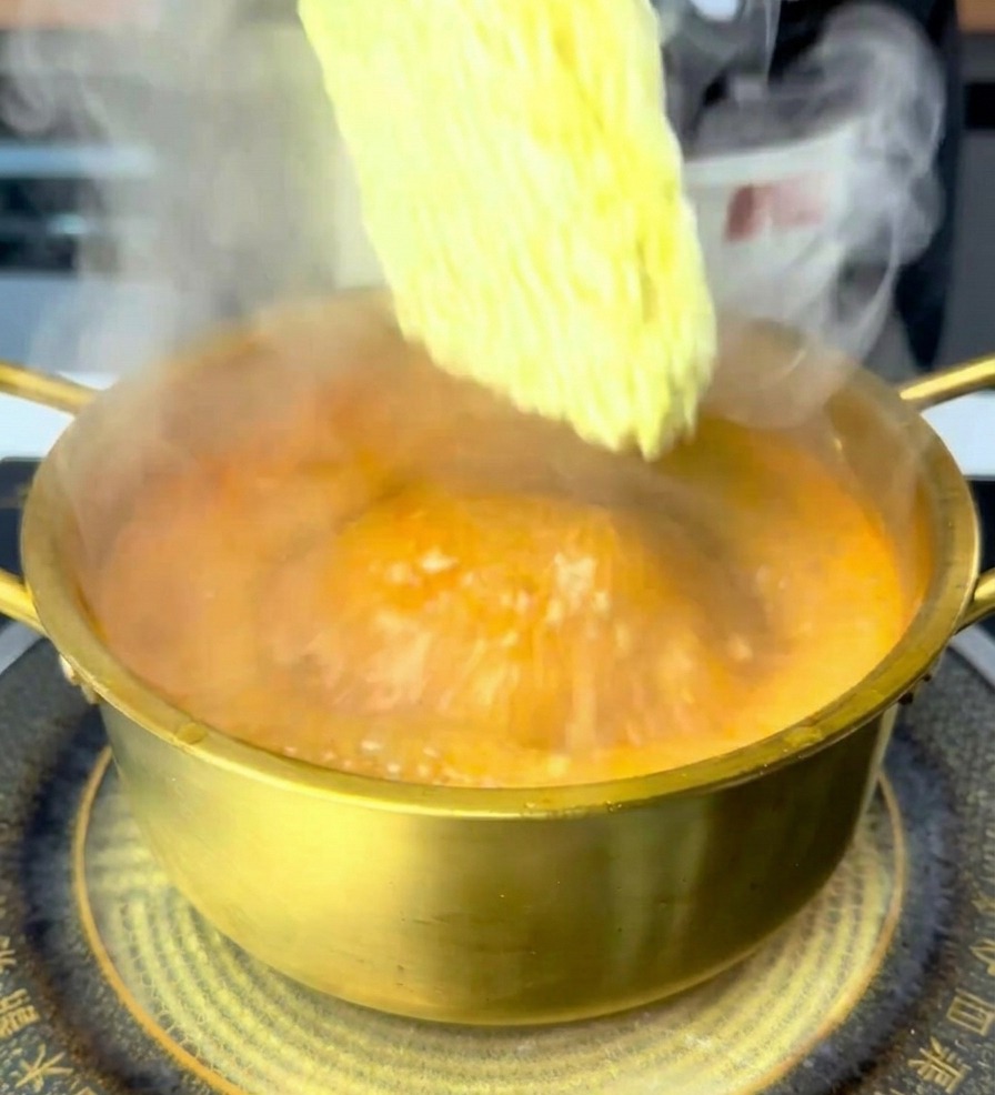 A hand placing a square block of yellow dried ramen noodles into a golden pot filled with steaming, bubbling orange-red spicy broth.