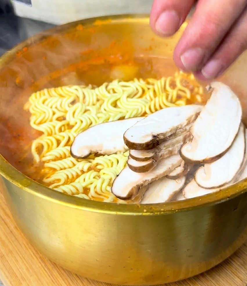 A hand placing sliced shiitake mushrooms onto a block of wavy instant noodles submerged in boiling red soup.