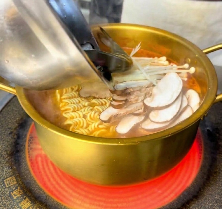 Fresh shrimp and mussels being poured from a stainless steel bowl into a golden pot containing boiling noodles and mushrooms.