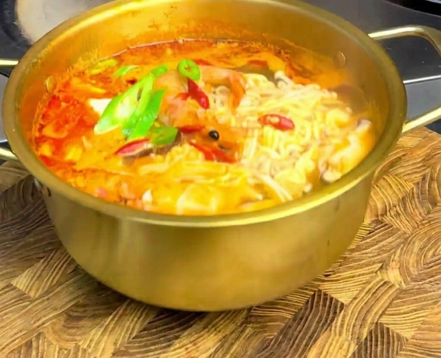 Spicy seafood ramen in a golden pot topped with fresh green and red chili slices, sitting on a rustic wooden cutting board.