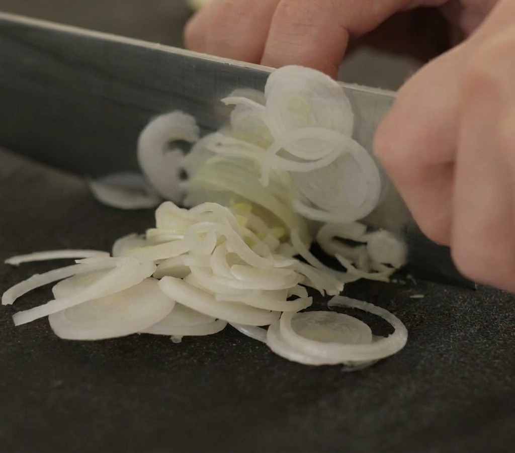 A close-up shot of hands using a sharp chefs knife to slice white leek stalks into thin rings on a black cutting board.