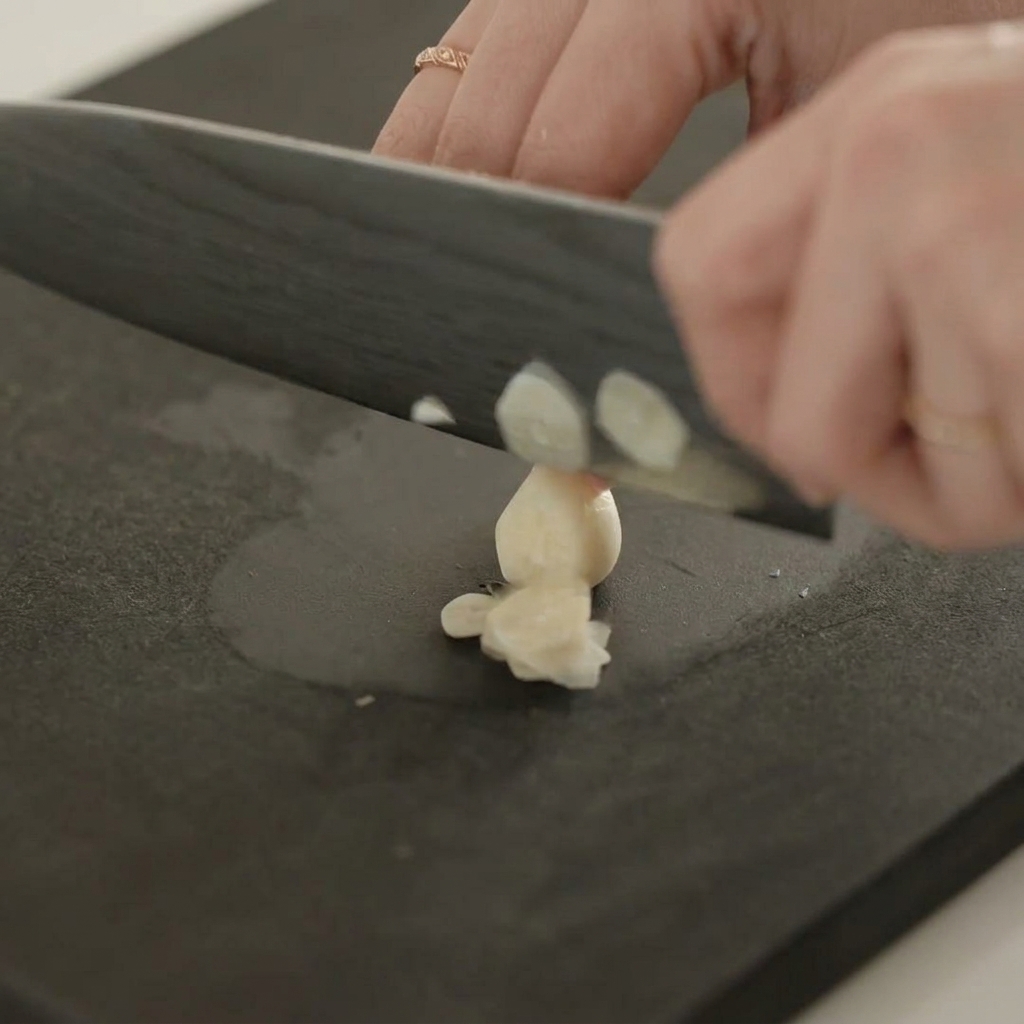 A close-up view of a hand using a sharp knife to thinly slice a garlic clove on a black cutting board.