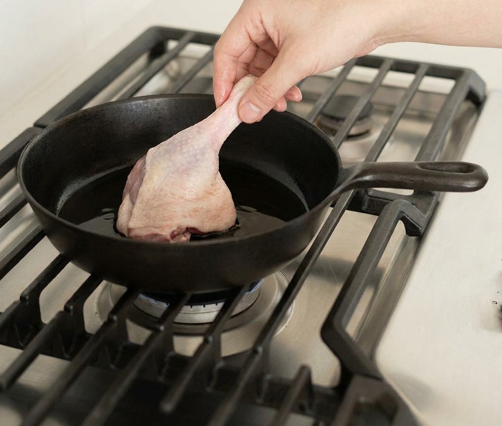 A hand placing a raw duck leg into a black cast iron skillet on a gas stove burner.