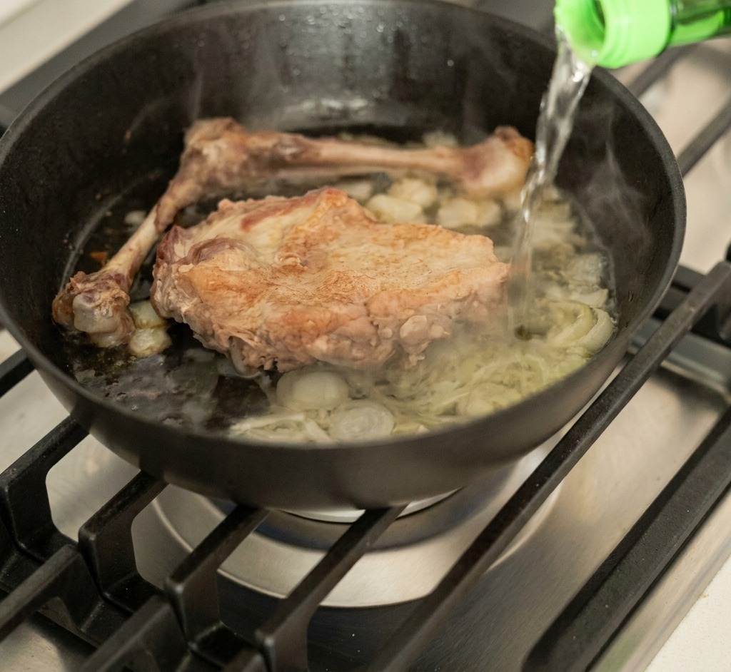 Liquid being poured from a green glass bottle into a steaming pan containing duck and aromatics.