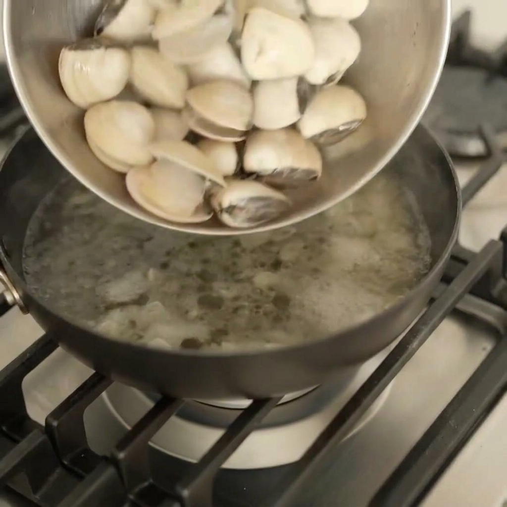 Fresh white clams being poured from a metal bowl into a dark pan on a gas stove.