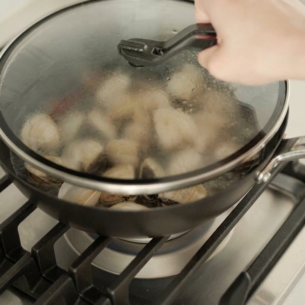 A hand placing a glass lid onto a pan filled with clams to steam them over a gas burner.