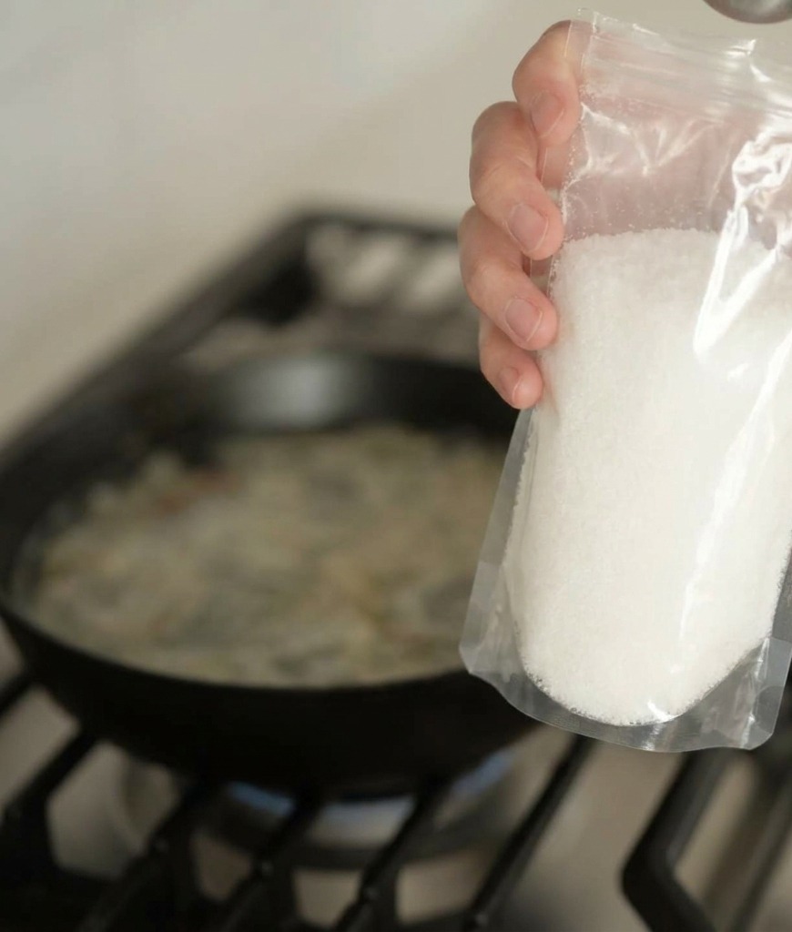 A hand holding a clear plastic bag and pouring white seasoning powder into a pan of simmering broth.