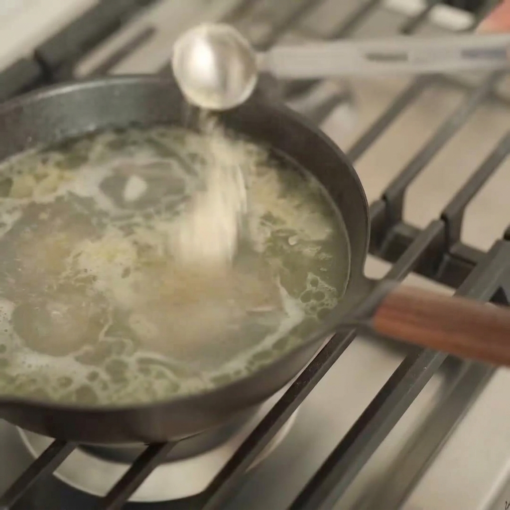 A silver measuring spoon pouring a dark liquid into a pot of bubbling light-colored ramen broth.