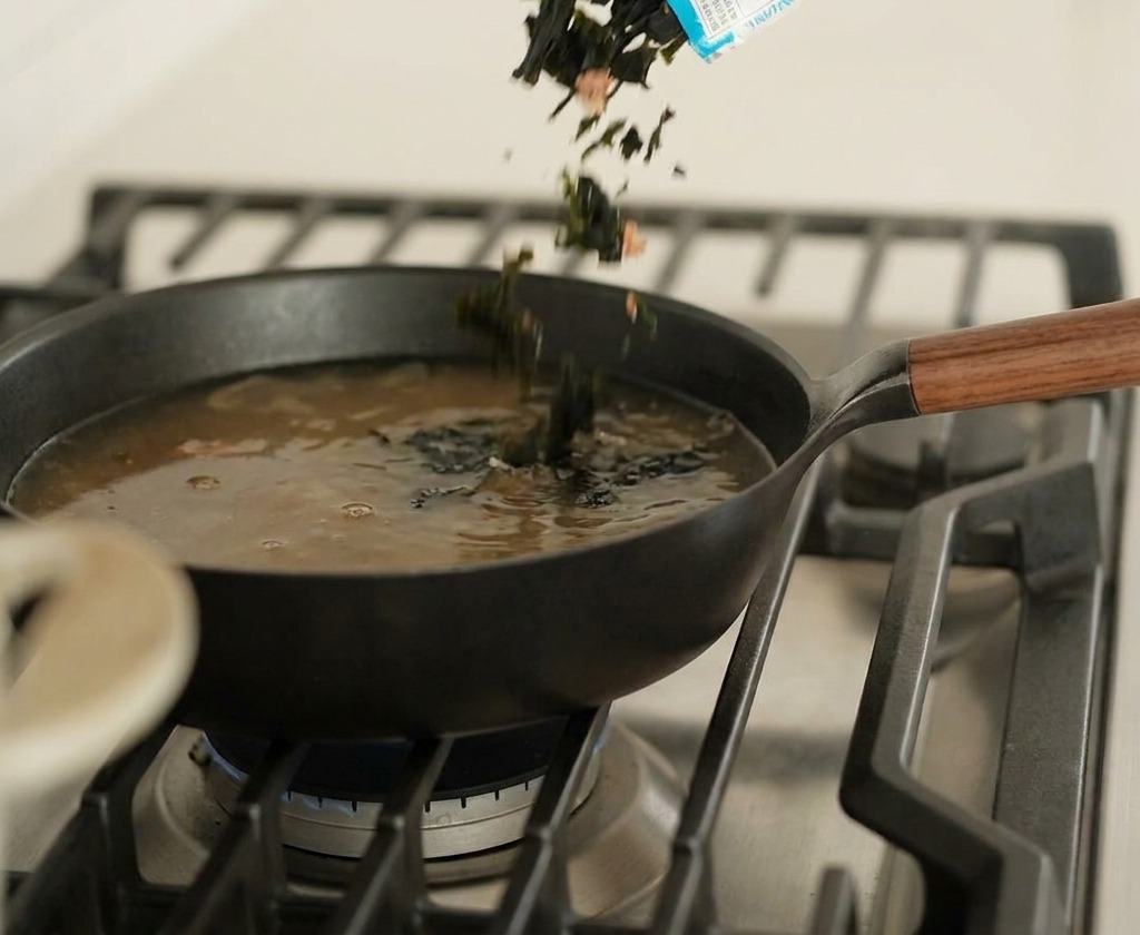 Dried green seaweed pieces being poured from a blue and white packaging into a black pan containing simmering dark broth.