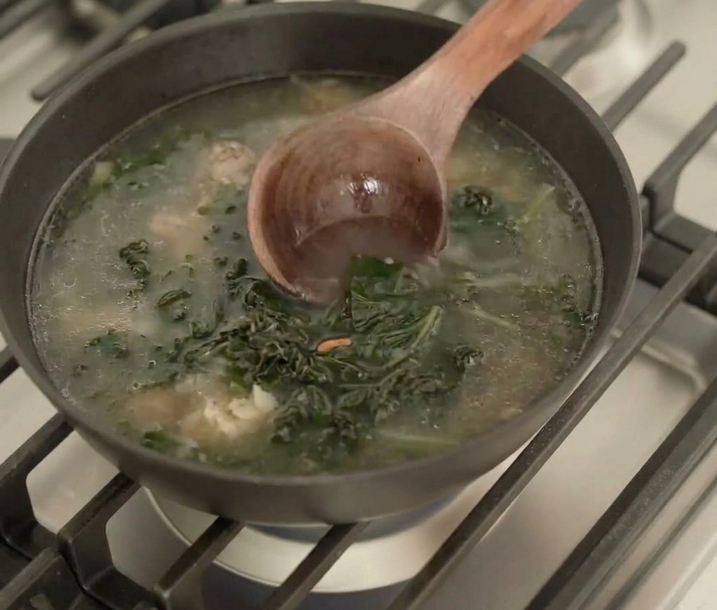 A wooden ladle stirring a dark, simmering broth containing rehydrated seaweed pieces in a black pan on a stovetop.