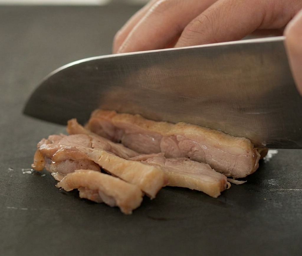 A sharp knife slicing through a piece of cooked, browned duck meat on a dark cutting board.