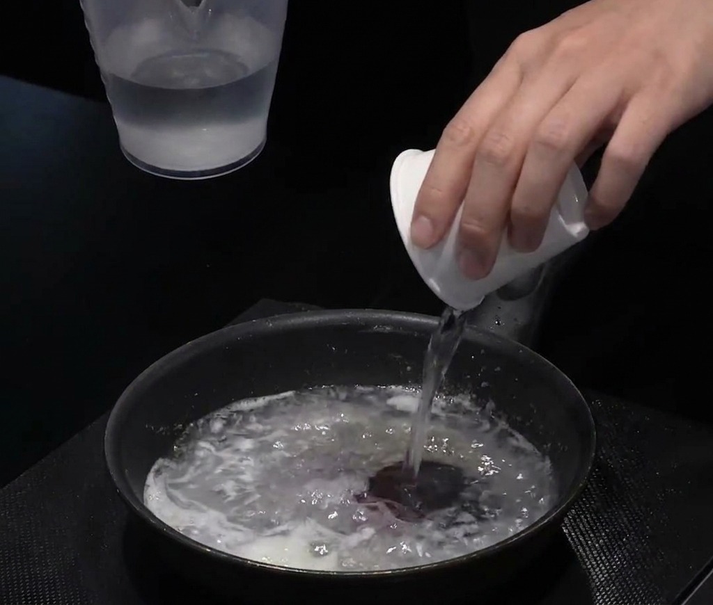 Water being poured from a small white cup into a black pan containing an oil and flour mixture to create a liquid base.