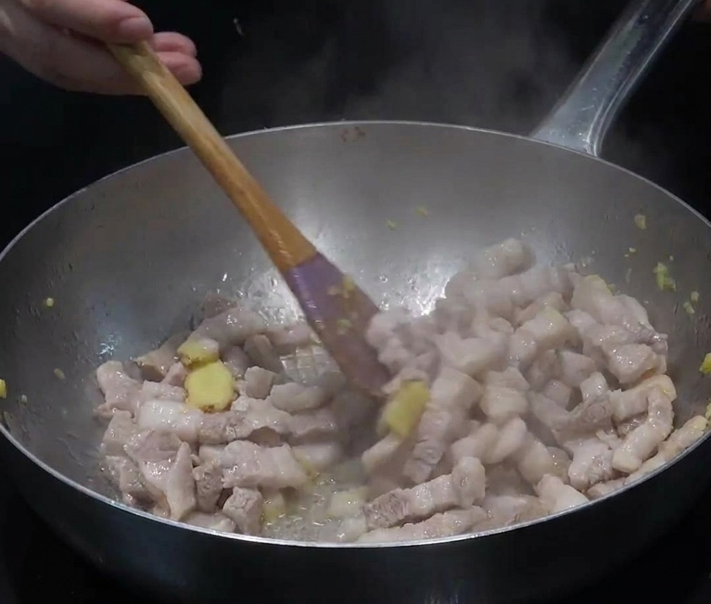 Slices of pork belly and minced ginger being stir-fried in a large silver stainless steel pan with steam rising.