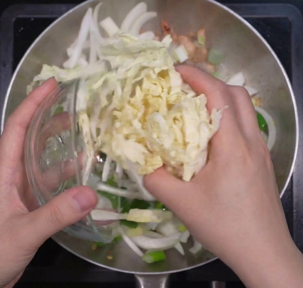 Hands adding a large portion of shredded napa cabbage into a pan containing sautéed onions and green onions.