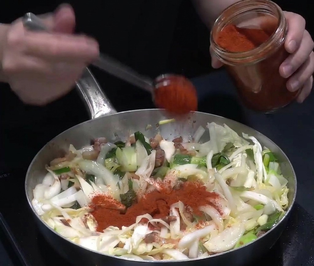 Bright red chili powder being spooned from a glass jar onto a mixture of sautéed vegetables and pork in a large pan.