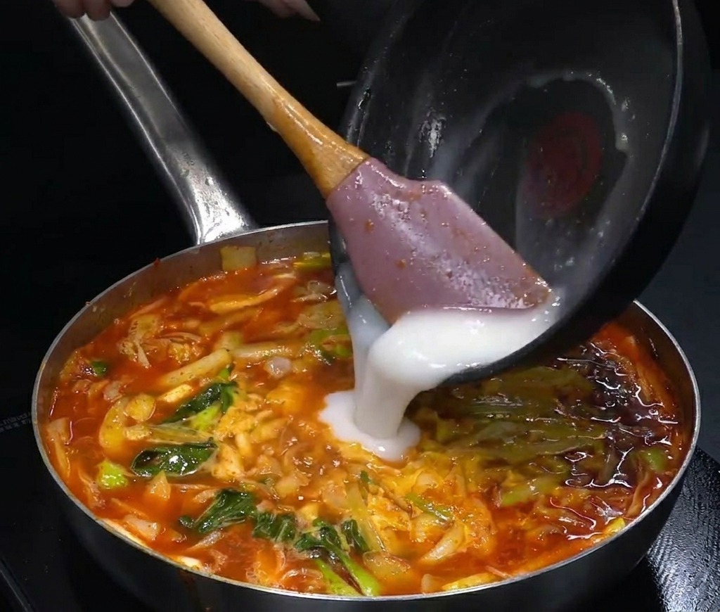 A white opaque liquid being poured from a small black pan into a large pot of bubbling red spicy soup.
