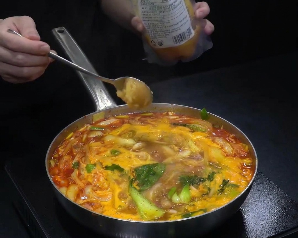 A spoonful of minced garlic being added to a wide pan of boiling red soup filled with cabbage and onions.