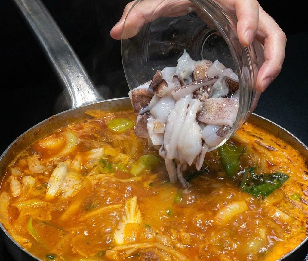 Raw sliced squid pieces being poured from a clear glass bowl into a steaming pot of red seafood broth.