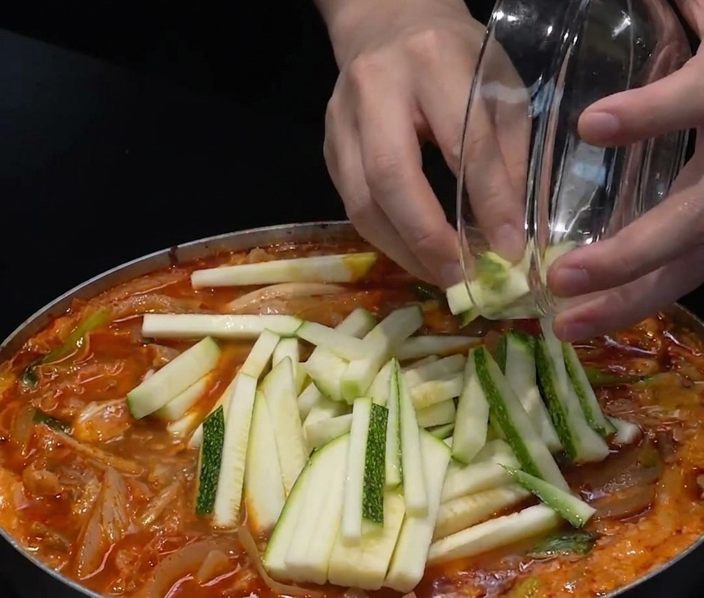 Sliced green-skinned zucchini and chopped green chili peppers being added to the top of a pot of red seafood soup.