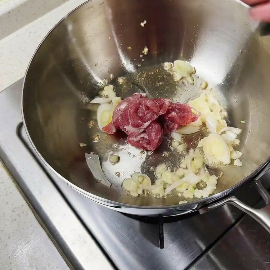 Pieces of raw pink pork being stir-fried with aromatics in the bottom of a metal pot.