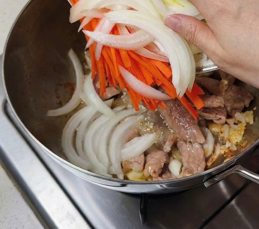 Hand pouring a plate of sliced onions, orange carrots, and green zucchini into the pot with the meat.