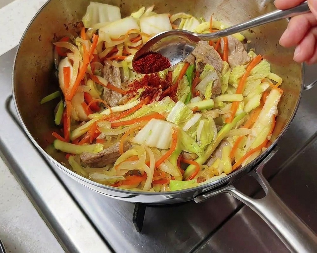 Vibrant red Korean chili powder being sprinkled from a spoon onto the stir-fried vegetables in the pot.