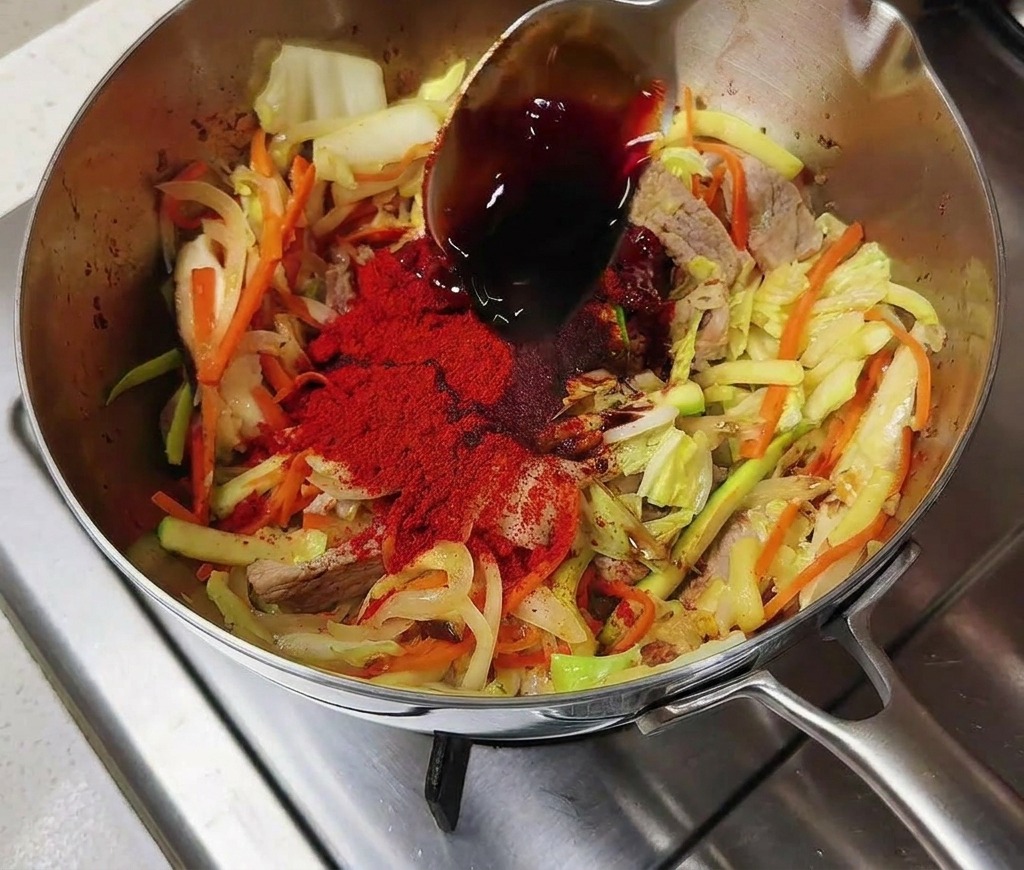 A silver spoon pouring dark soy sauce onto a vibrant pile of red chili powder and stir-fried vegetables in a stainless steel pot.