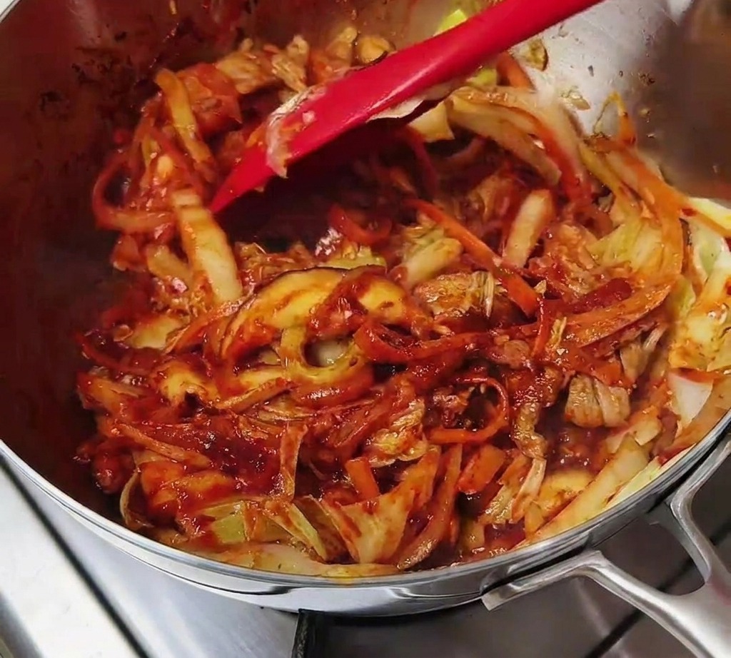 A red spatula stir-frying a mixture of meat and vegetables that have been completely coated in a rich, red chili seasoning.