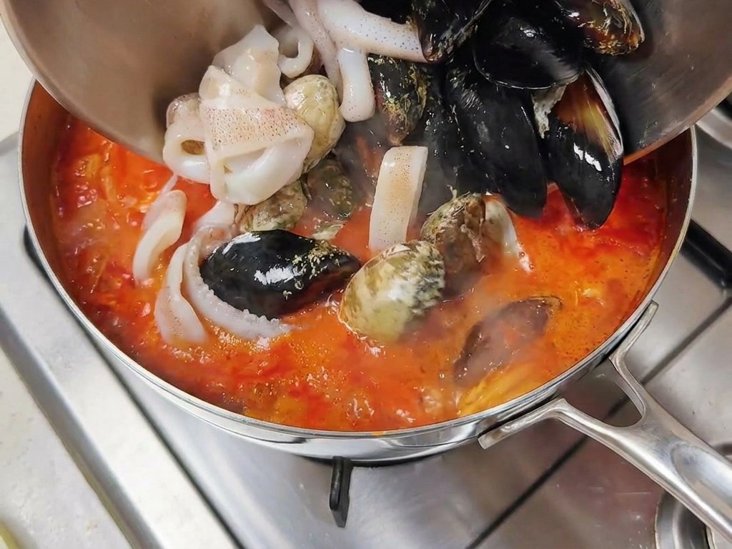 A variety of fresh seafood, including mussels in black shells and white squid rings, being poured from a bowl into a bubbling red broth.