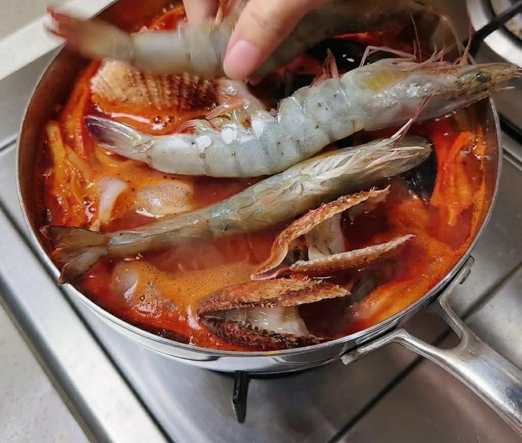 A hand placing several large raw gray shrimp into a pot of bubbling red spicy soup filled with various shellfish.