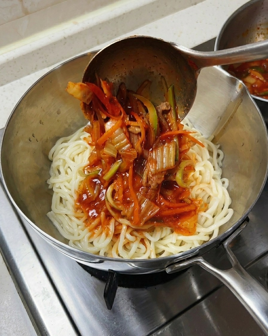 A metal ladle pouring spicy red seafood soup and vegetables over a mound of thick white noodles in a stainless steel bowl.