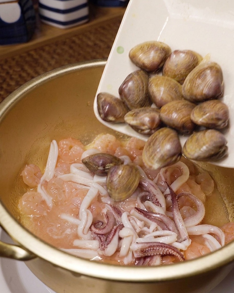 Fresh whole clams being poured from a white rectangular plate into a gold cooking pot containing stir-fried pink shrimp and squid rings.