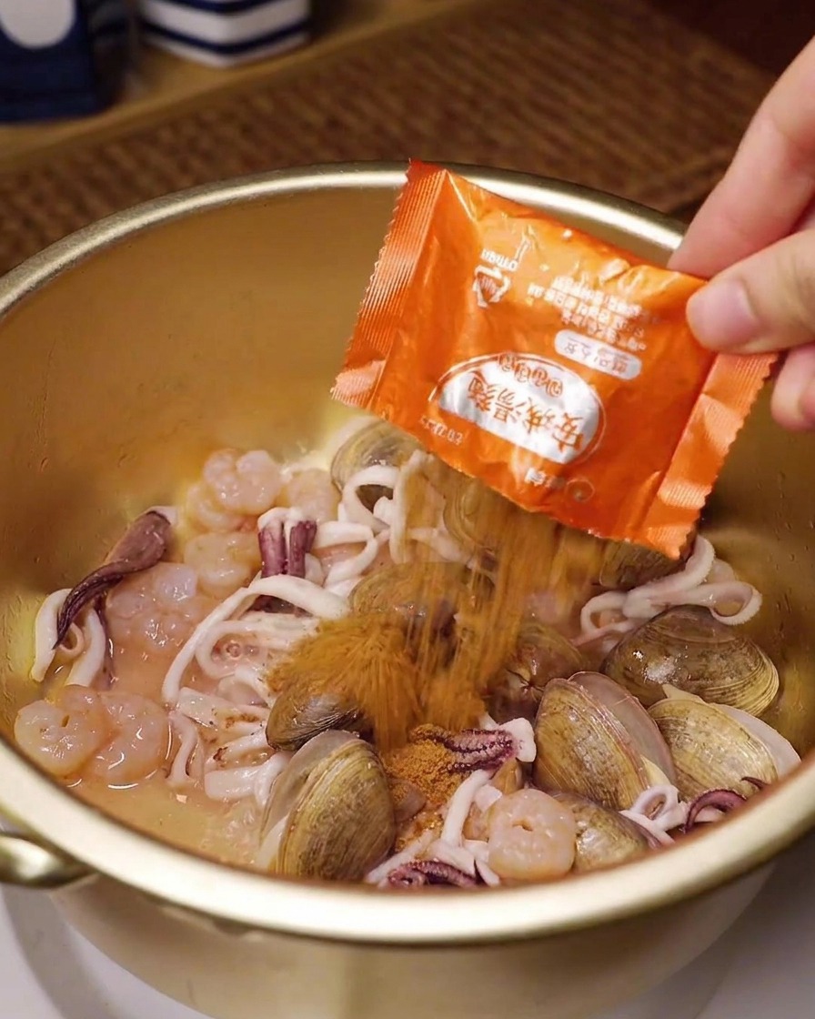 A hand pouring an orange packet of instant ramen seasoning over clams, squid, and shrimp in a golden brass pot.