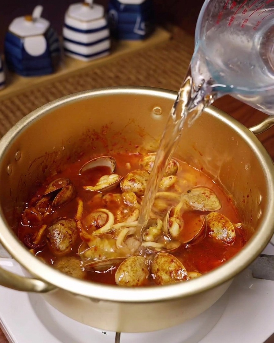 Fresh water being poured from a glass measuring pitcher into a golden pot containing the spicy stir-fried seafood.