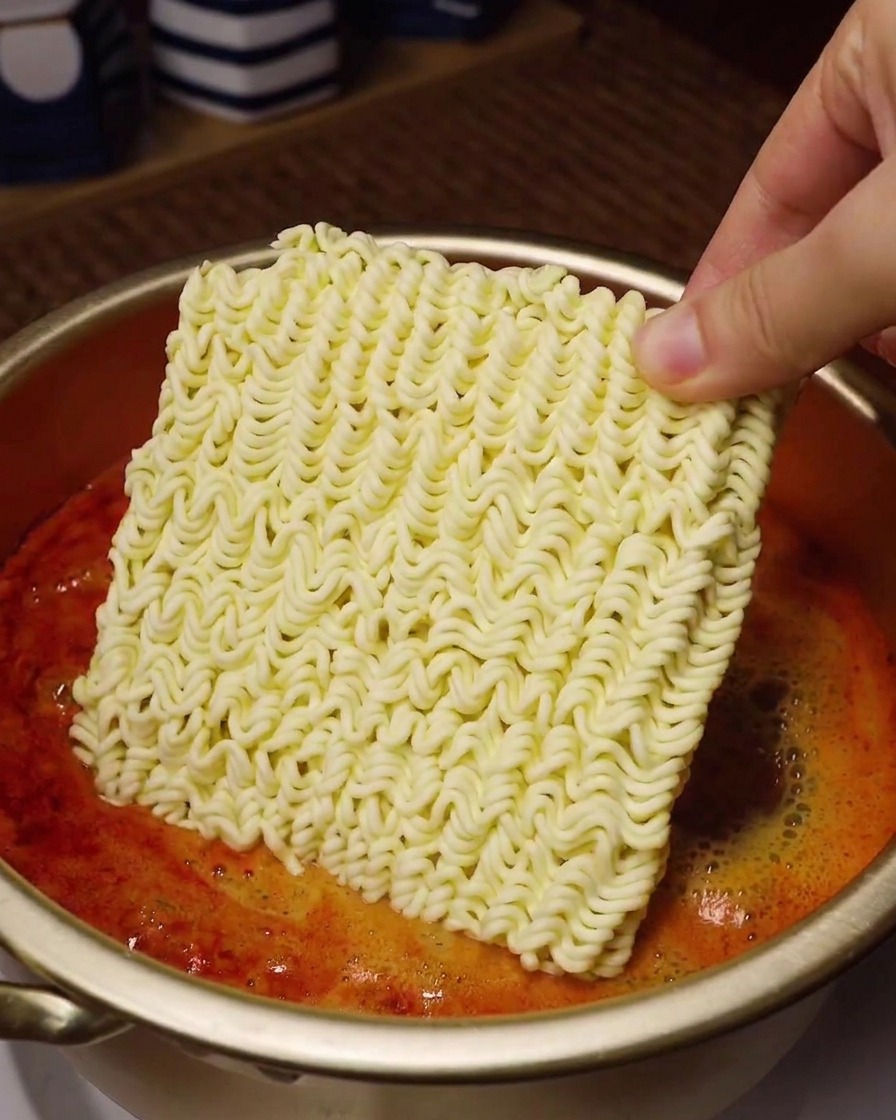 A hand holding a square block of dry instant noodles over a gold pot filled with bubbling, bright red spicy soup.