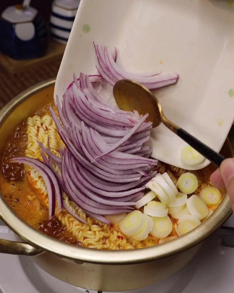 Thinly sliced purple red onions and round white scallion slices being scooped from a white plate into a pot of simmering ramen noodles in red broth.
