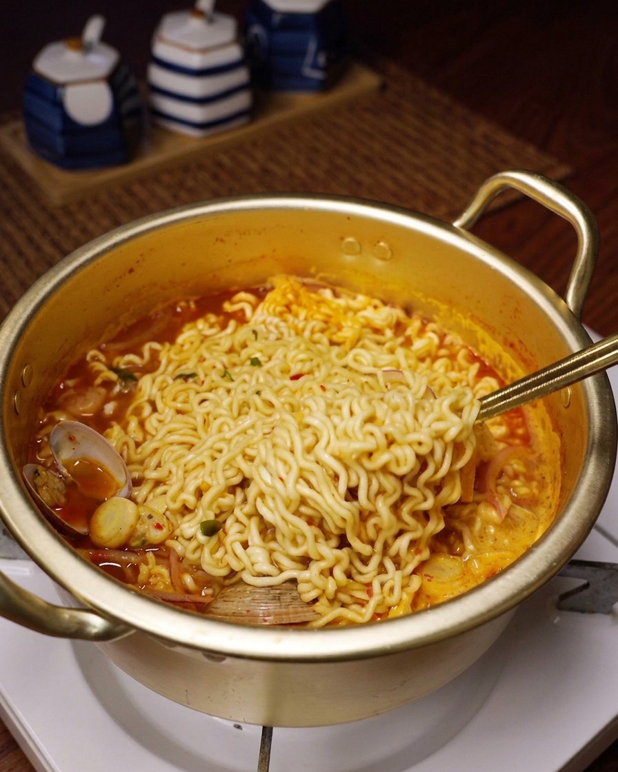 Wavy instant noodles being lifted out of a simmering red seafood broth with a metal spoon.