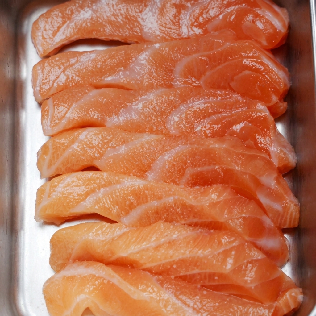 Slices of fresh raw salmon arranged in a metal tray, sprinkled with fine salt and black pepper granules.
