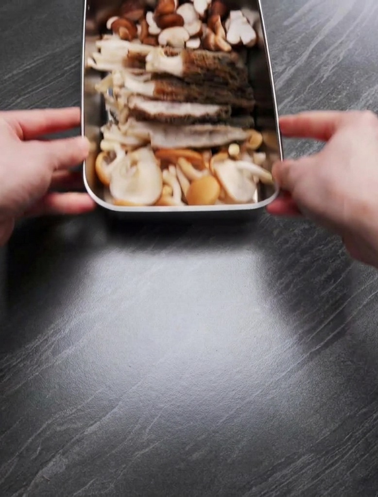 Hands holding a metal tray containing a mix of sliced morels and halved shiitake mushrooms on a dark countertop.