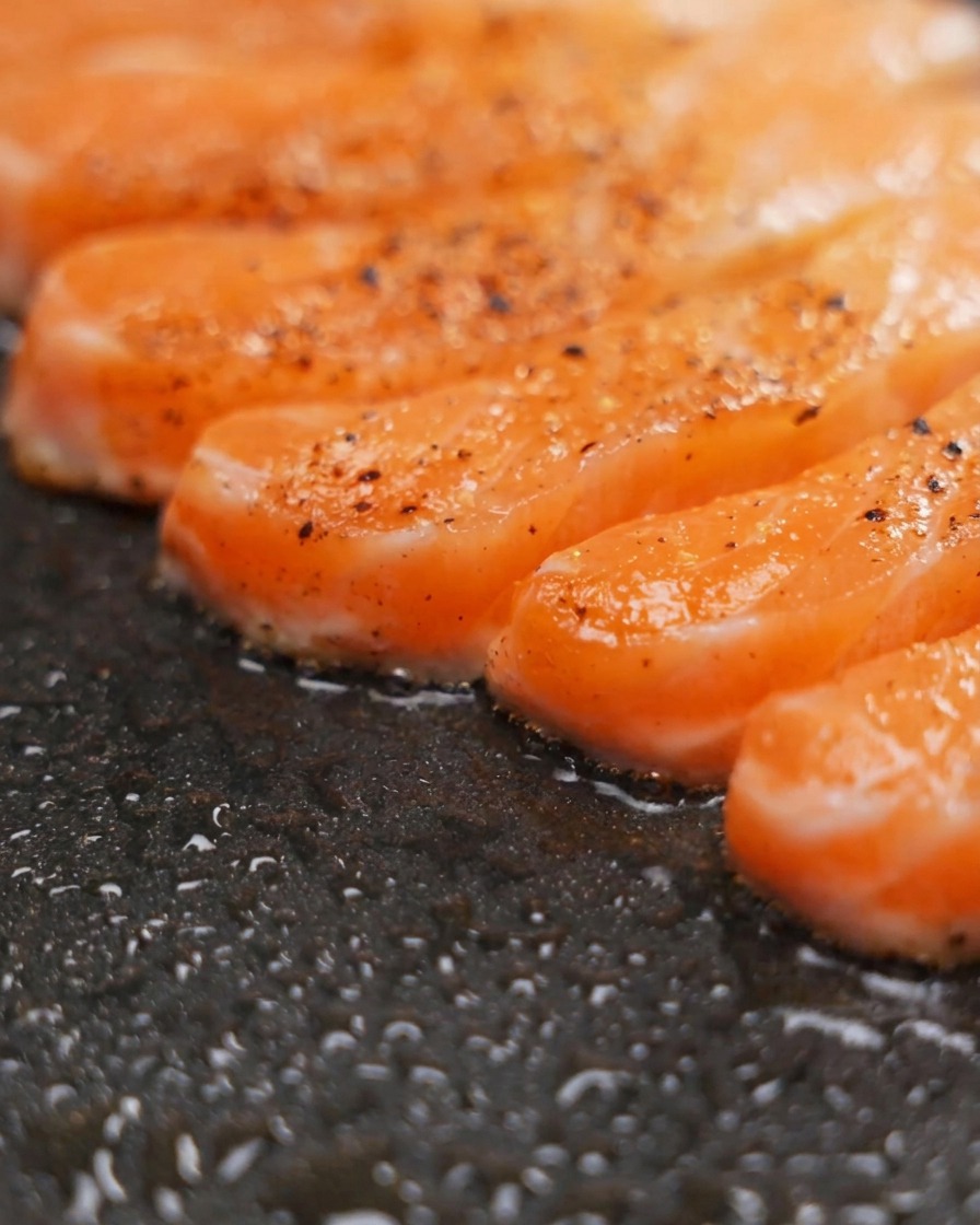 Seasoned salmon fillets sizzling in a dark non-stick pan, showing visible browning and pepper seasoning on the surface.