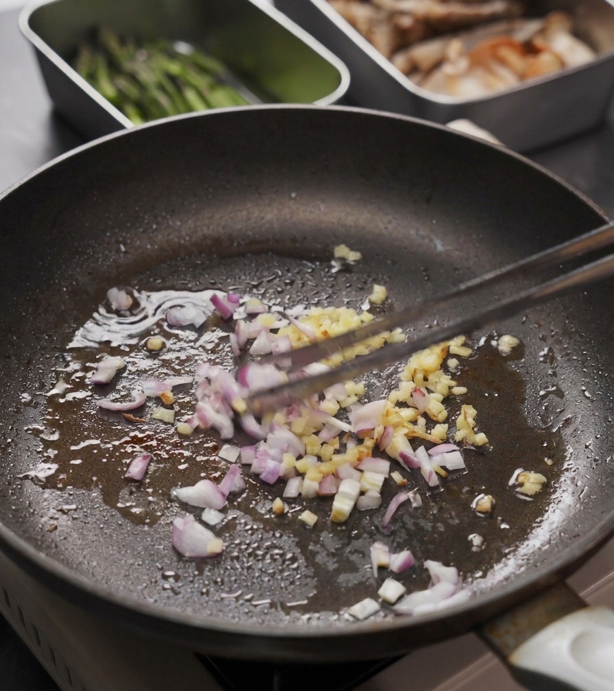 Minced ginger and red onions being stirred in a skillet with a pair of cooking tweezers.