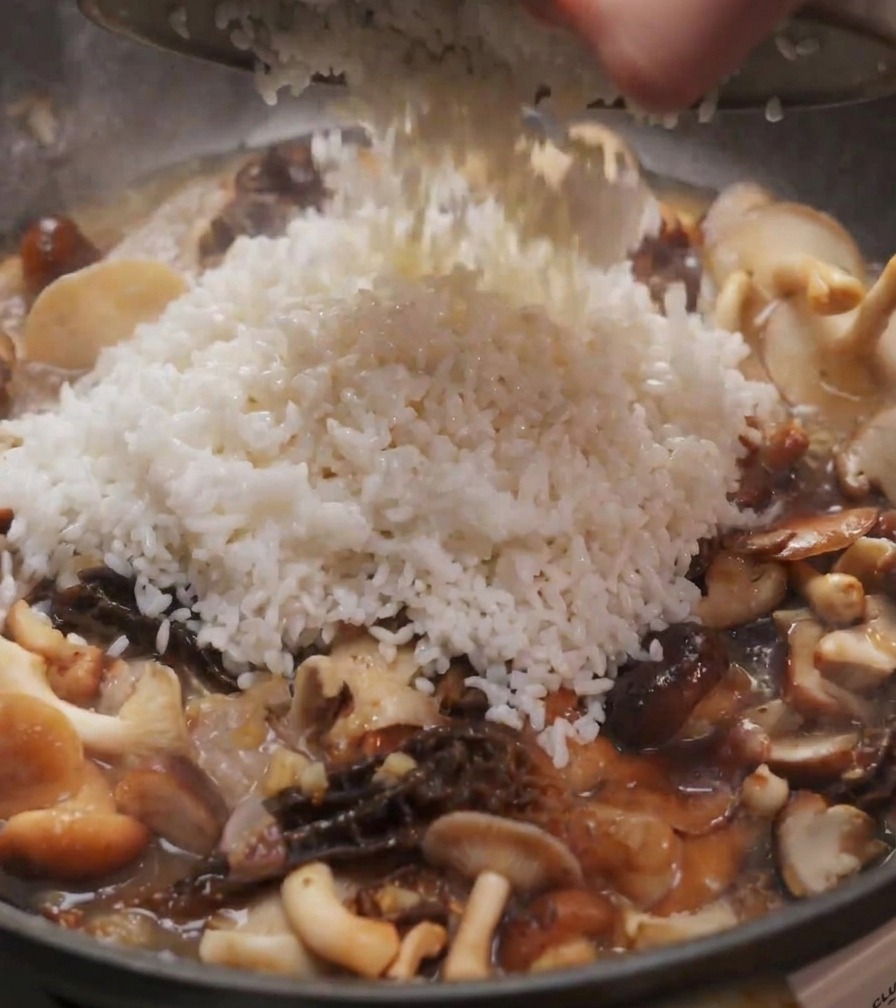 A heap of white rice being poured from a bowl into a pan filled with sautéed shiitake and morel mushrooms.