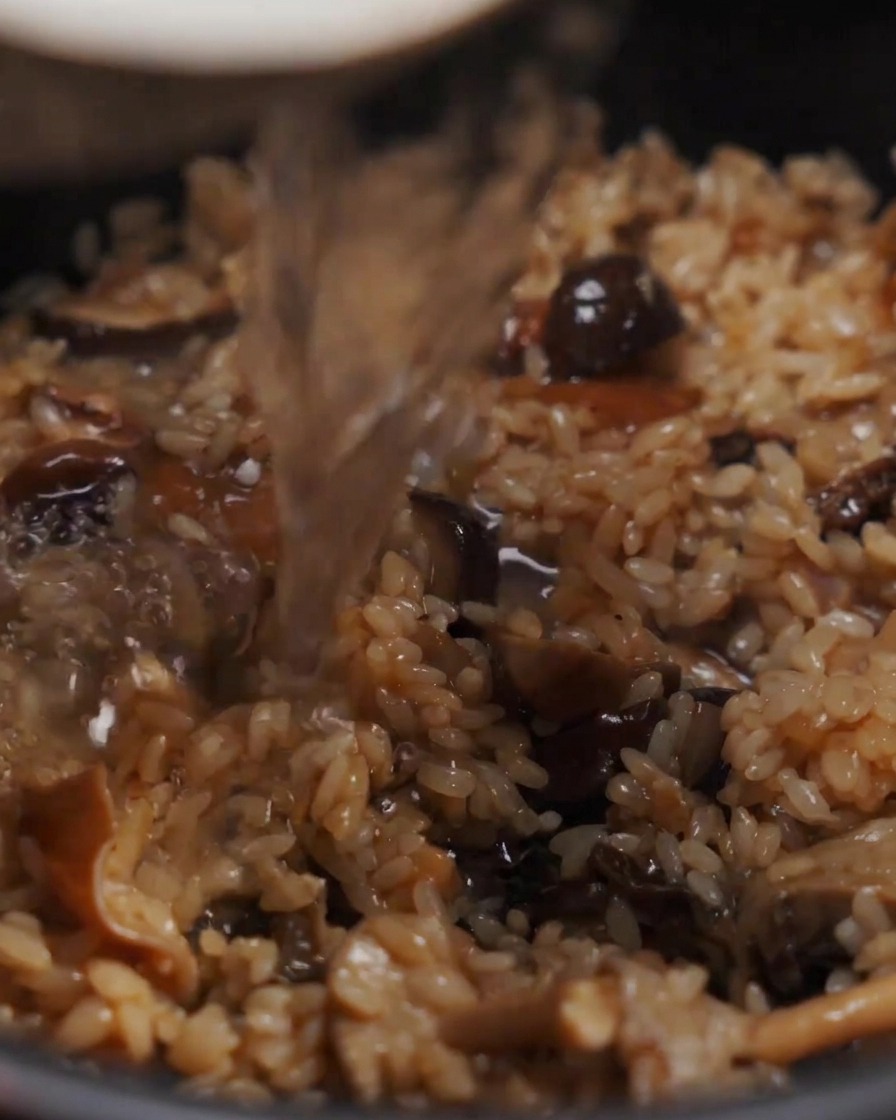 Water being poured into a pot containing a seasoned mixture of rice and mushrooms.