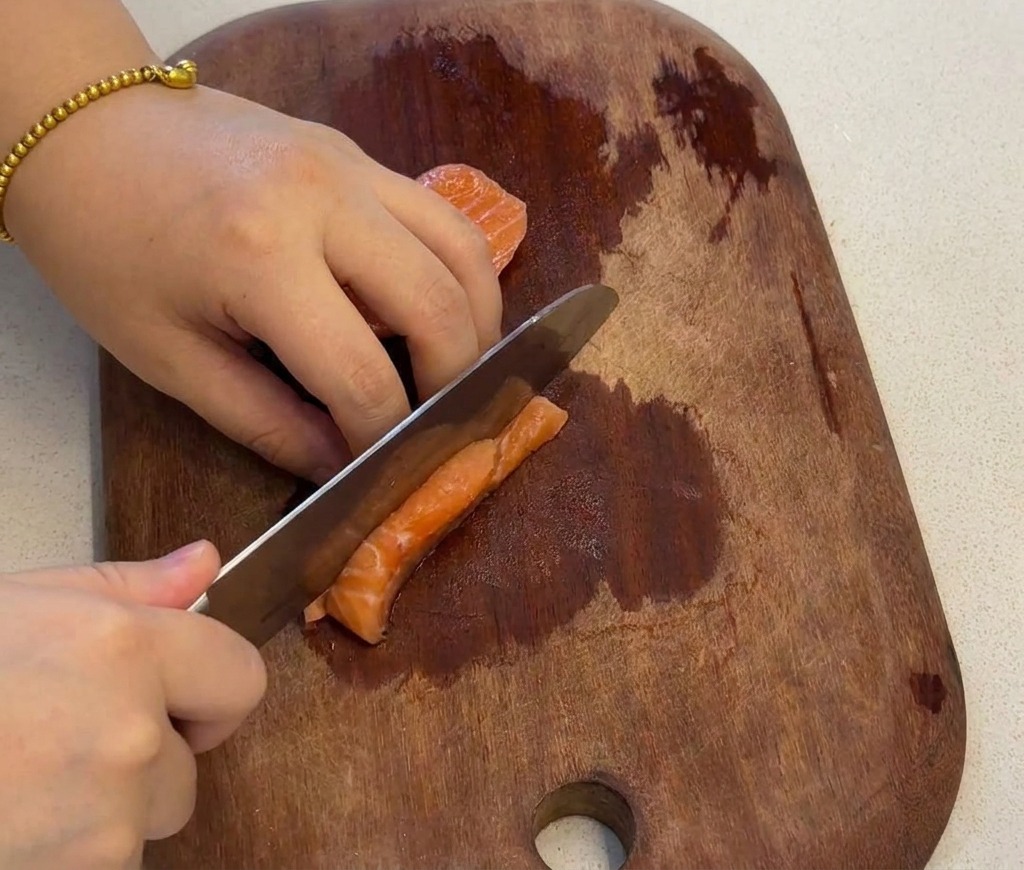 A hand using a knife to slice a fresh orange salmon fillet into small uniform cubes on a dark wooden cutting board.