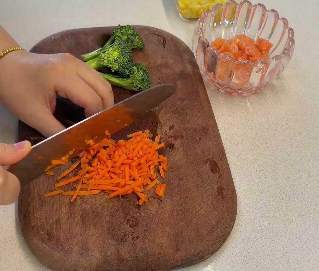 Shredded orange carrots being finely chopped with a chefs knife on a wooden board next to some prepared broccoli florets.